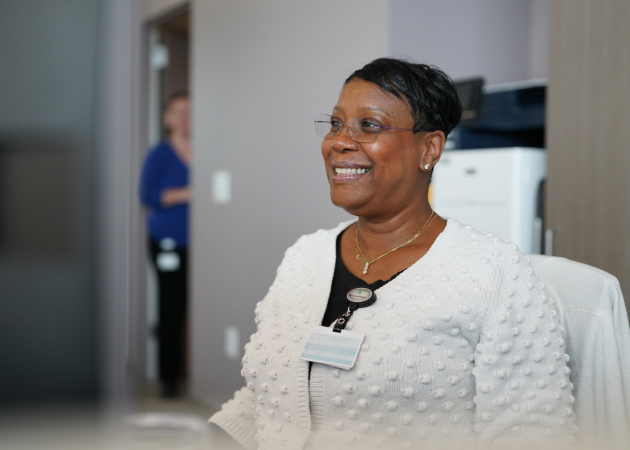 A woman wearing glasses and a white sweater sits at a desk, smiling. She has a lanyard with an ID badge, and another person is visible in the blurred background near a doorway.