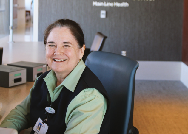 A woman with brown hair pulled back, wearing a light green shirt and dark vest, sits and smiles at a desk in a professional office setting with "Main Line Health" visible on the wall behind her.