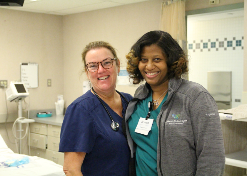 Two smiling healthcare professionals stand together in a medical room; one wears navy scrubs and glasses, the other has a gray jacket and a name badge. Medical equipment and a hospital bed are visible in the background.