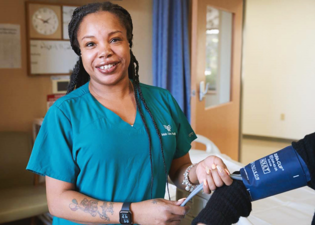 A smiling healthcare worker in green scrubs takes a patient’s blood pressure in a hospital room, holding a blood pressure cuff and stethoscope.