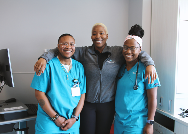 Three smiling people stand together in a medical office. Two wear teal scrubs, one with a stethoscope, and the person in the middle wears a gray jacket. They look happy and friendly, with arms around each other.