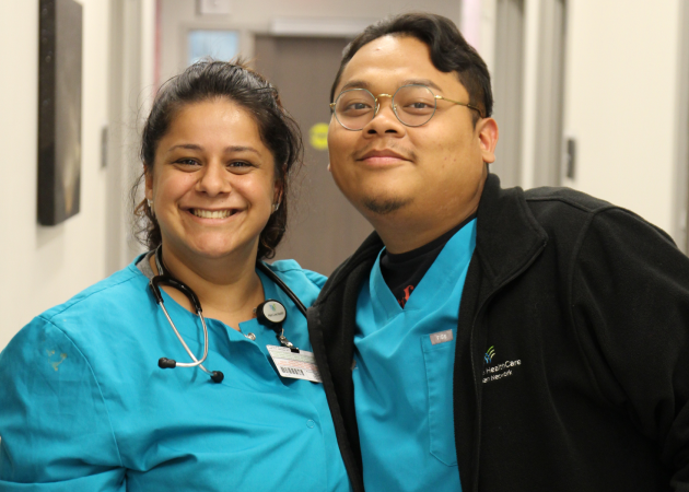 Two healthcare professionals in blue scrubs and badges smile at the camera in a hallway. One wears a stethoscope around her neck; the other wears glasses and a black jacket.