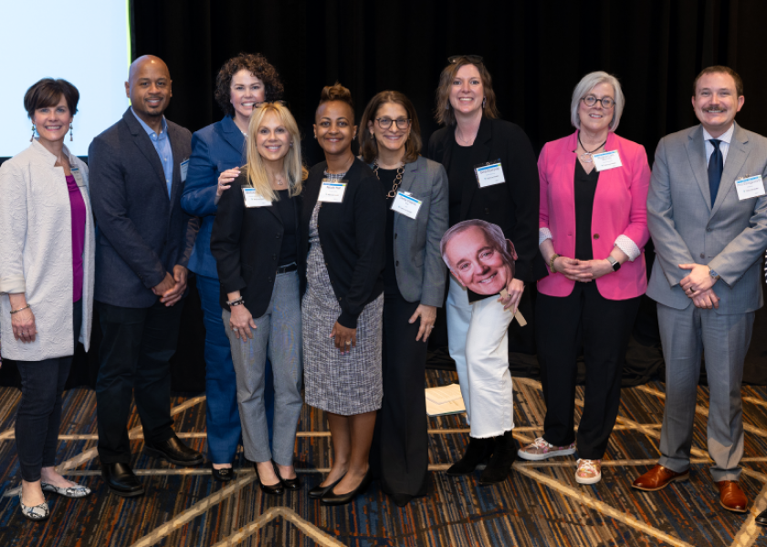 Nine professionally dressed people pose and smile for a group photo indoors; one person holds a large cutout of a man's face. The group stands on a carpet with geometric patterns, in front of a black curtain.