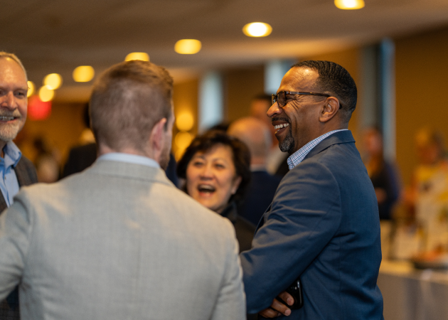 A group of professionally dressed people stand together indoors, smiling and laughing as they converse at what appears to be a networking event or conference. Warm lighting creates a friendly, welcoming atmosphere.