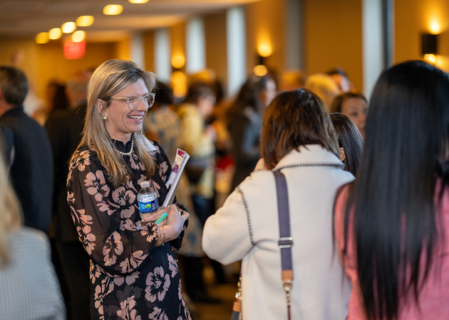 A woman in a floral dress, holding a water bottle and folder, smiles while speaking with others in a crowded, warmly-lit indoor event space. People are gathered in small groups, talking.