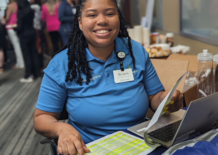 A smiling woman in a blue polo shirt sits at a table with a laptop and highlighted documents. She is wearing a name badge and is interacting with people at an event or office setting.