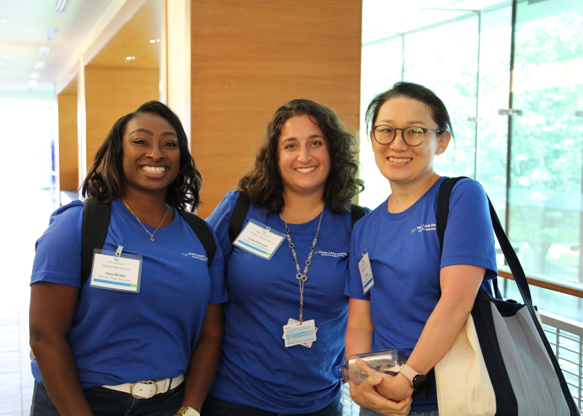 Three women wearing blue shirts and name tags stand together indoors, smiling at the camera. Two carry tote bags, and one holds a phone. They appear to be at a professional or academic event.