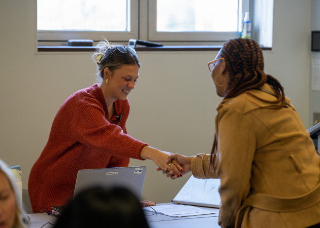 Two women smile and shake hands across a desk in an office setting. One sits with a laptop open, while the other stands, both appearing friendly and engaged.