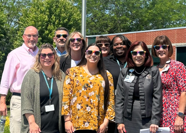 A group of nine adults, most wearing sunglasses with rainbow stripes, smiles outdoors in front of a brick building and trees. Several wear ID badges, and the weather appears sunny.