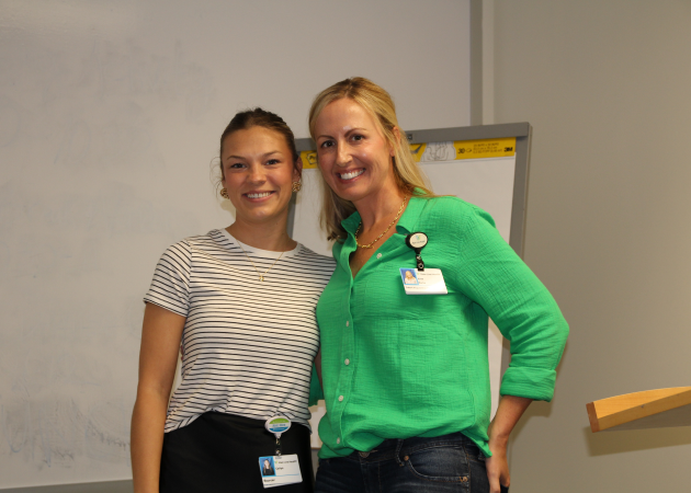 Two women wearing work badges smile and stand together in front of a whiteboard and a flip chart in an office or classroom setting.