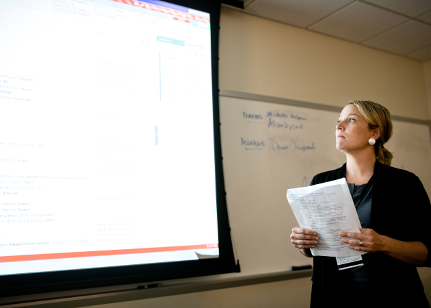 A woman holding papers stands in front of a projected screen and a whiteboard, looking thoughtfully at the screen during a presentation or lecture in a classroom.