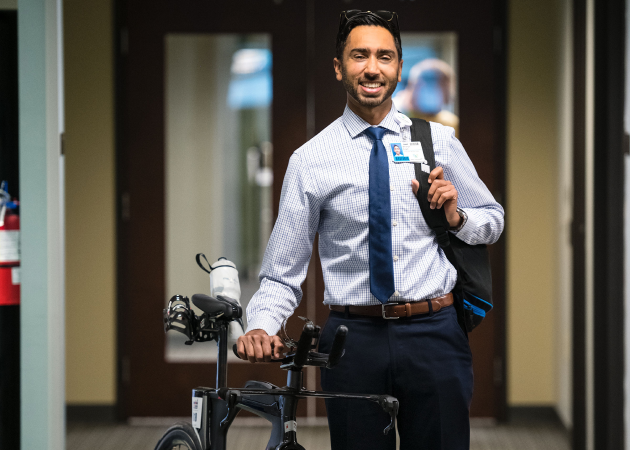 A man in business attire smiles while holding a bicycle and carrying a shoulder bag, standing in an office hallway with double doors in the background.