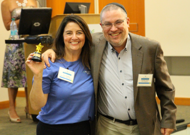 A smiling woman holds a small gold star trophy while standing next to a smiling man in a suit. They both wear name tags and are posing for a photo in a conference room.
