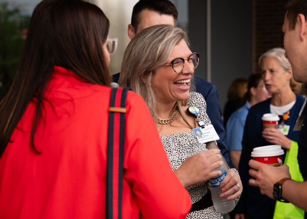 A woman with glasses smiles and holds a water bottle while talking with others outdoors. People around her wear work badges and are holding coffee cups, suggesting a casual work gathering.