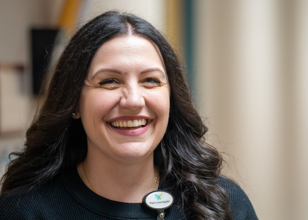 A woman with long dark hair wearing a black top and a name badge smiles brightly while standing indoors with a blurred background.
