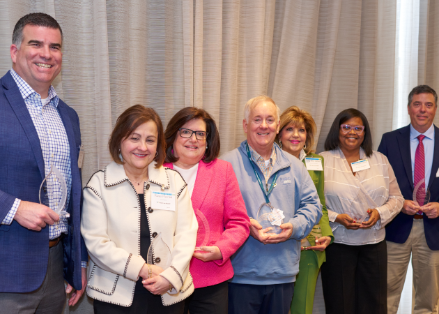 Seven people stand in a row indoors, smiling and holding glass awards. They are dressed in business or business casual attire and stand in front of beige curtains.