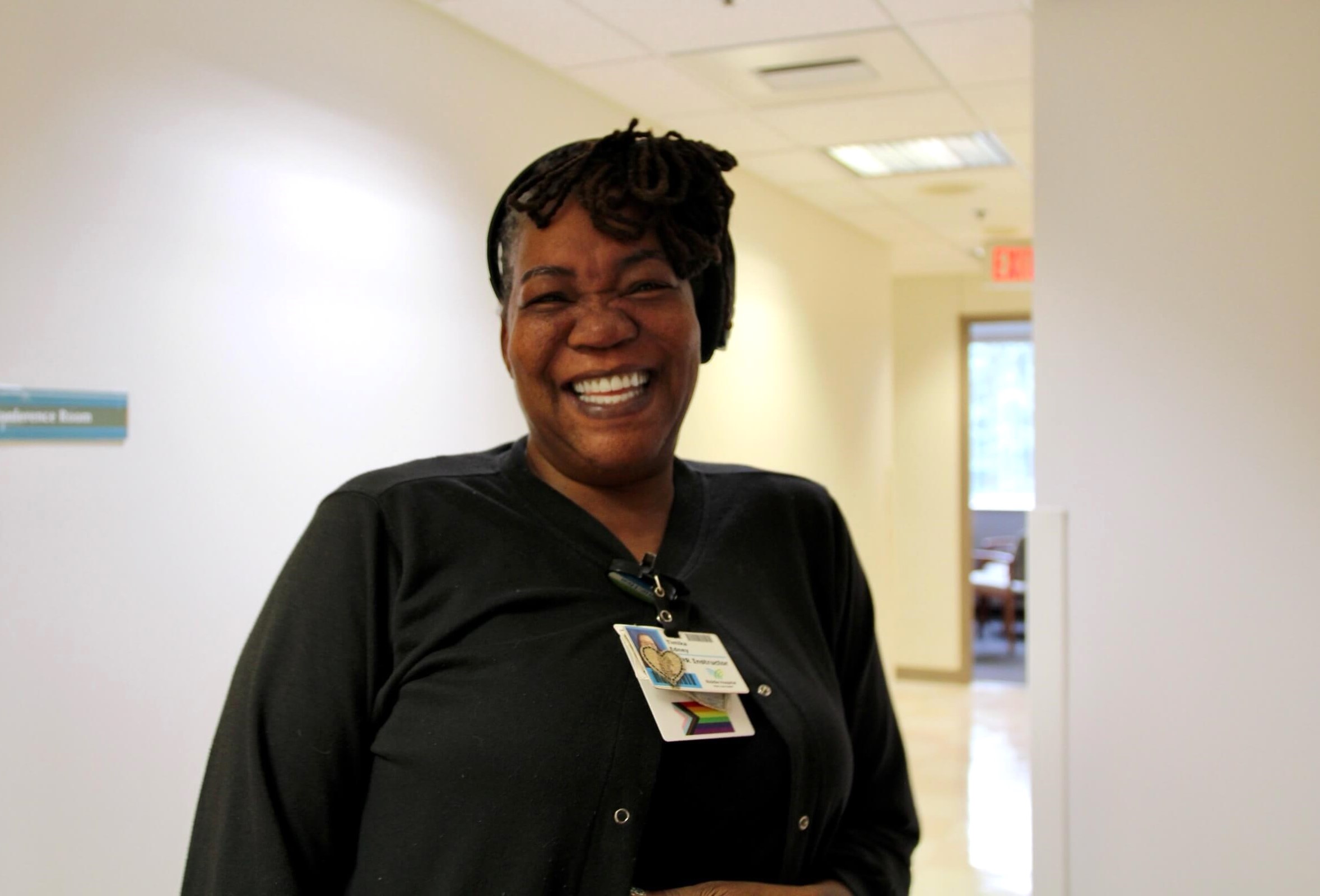 A woman wearing a black sweater and a headscarf smiles brightly in a hallway, displaying an ID badge clipped to her chest. The background is an indoor office setting with cream-colored walls.