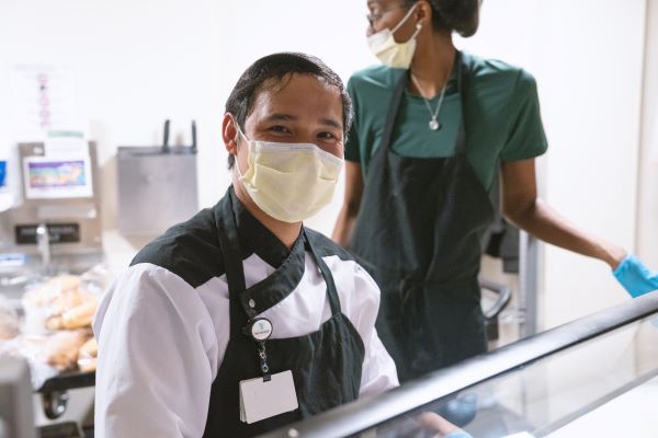 Two cafeteria workers wearing face masks and black aprons stand behind a counter. One, a man, smiles at the camera while the other, a woman, looks to the side. Food items are visible in the background.