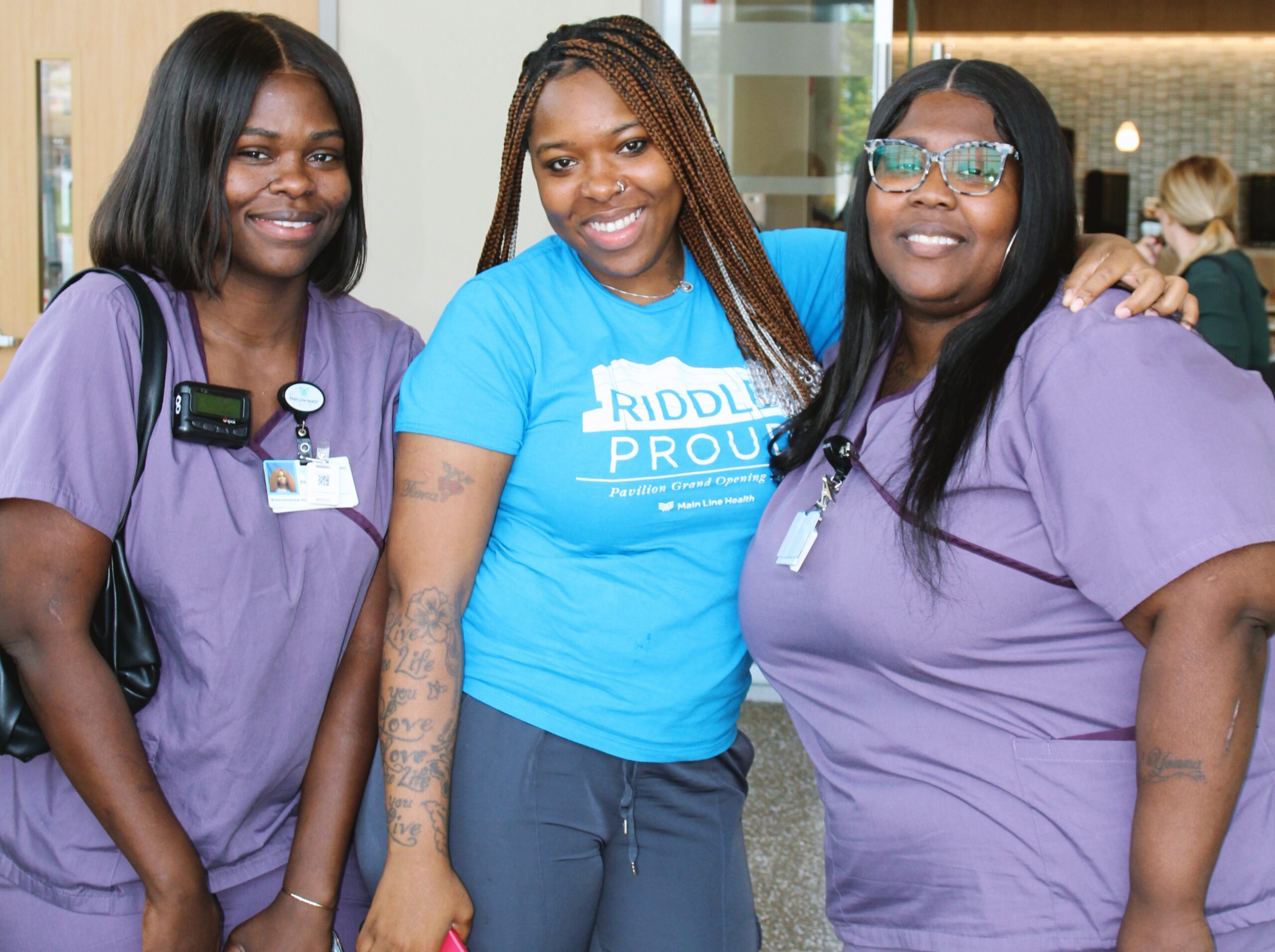 Three women smile and pose together indoors; two wear purple scrubs and ID badges, while the woman in the middle wears a blue “Riddle Proud” T-shirt. They appear happy and friendly in a bright, modern setting.