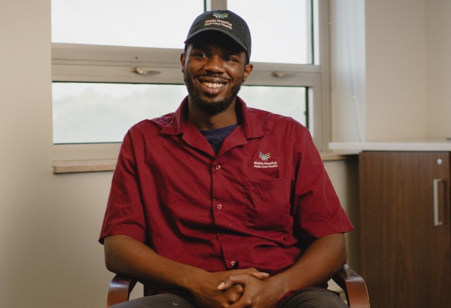 A man wearing a maroon short-sleeve shirt and a navy cap sits and smiles in an office chair, with a window and cabinet in the background.