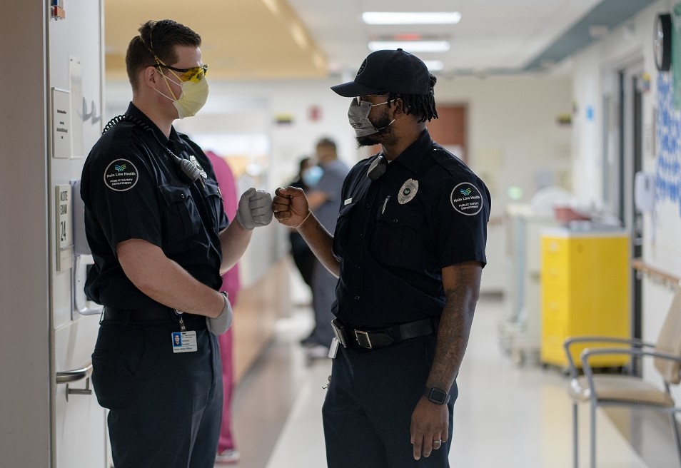 Two security officers in uniform, wearing face masks, greet each other with a fist bump in a hospital hallway. One officer is wearing gloves and goggles. The corridor is brightly lit, with medical staff in the background.