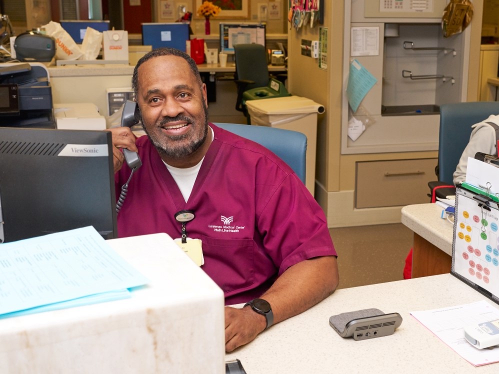 A smiling healthcare worker in burgundy scrubs sits at a desk, talking on the phone in a busy hospital or clinic reception area. Papers and office equipment are visible around him.