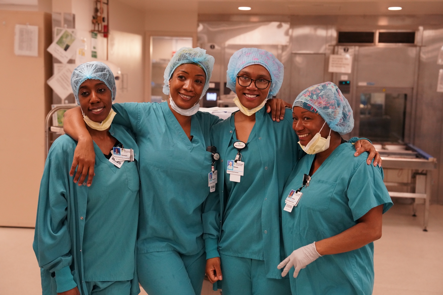 Four healthcare workers in teal scrubs and hairnets smile and pose together in a hospital setting, with one person’s arm around another. They stand in a brightly lit medical facility.