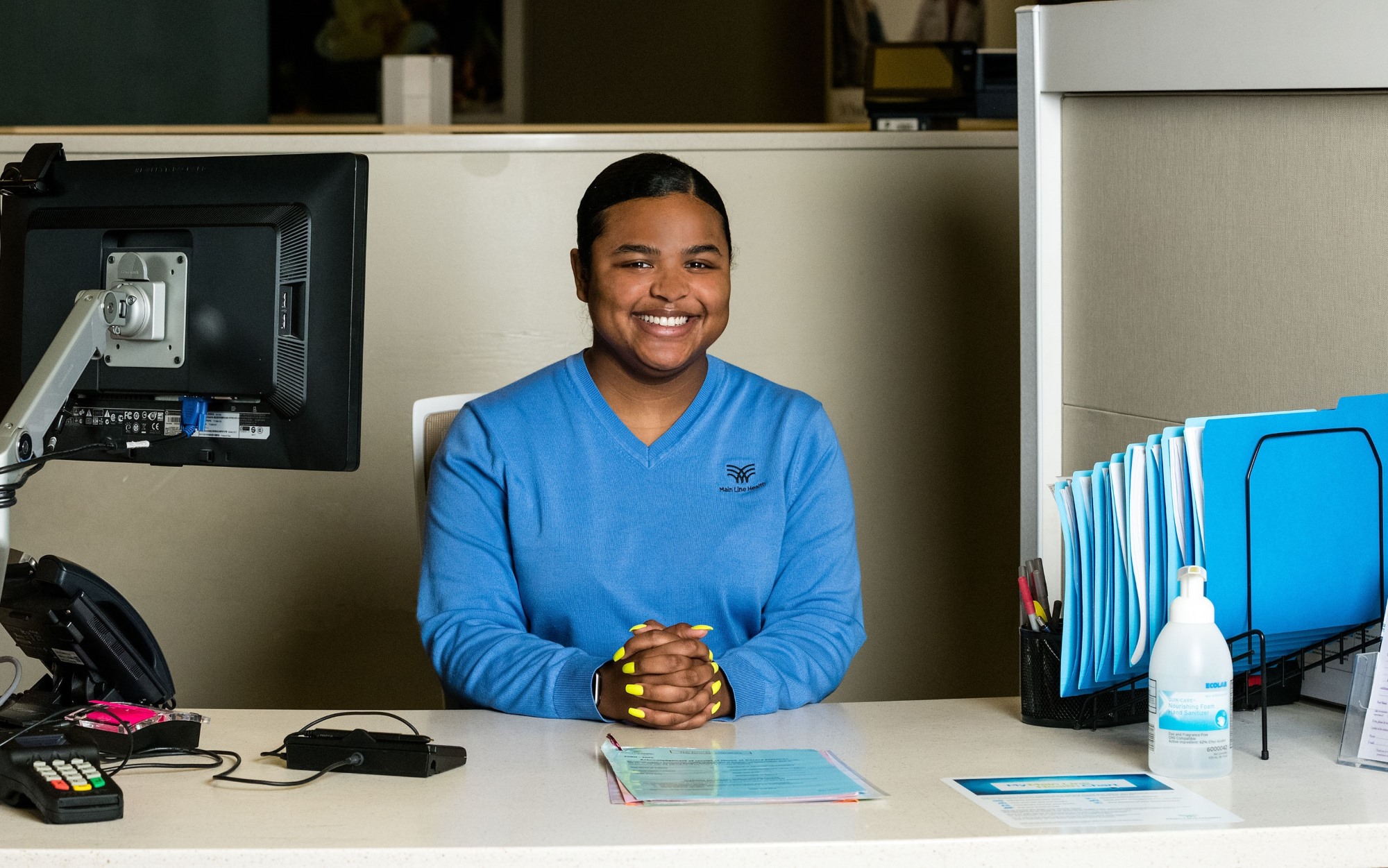 A smiling receptionist wearing a blue sweater sits at a desk with a computer monitor, paperwork, folders, and hand sanitizer in a professional office setting.