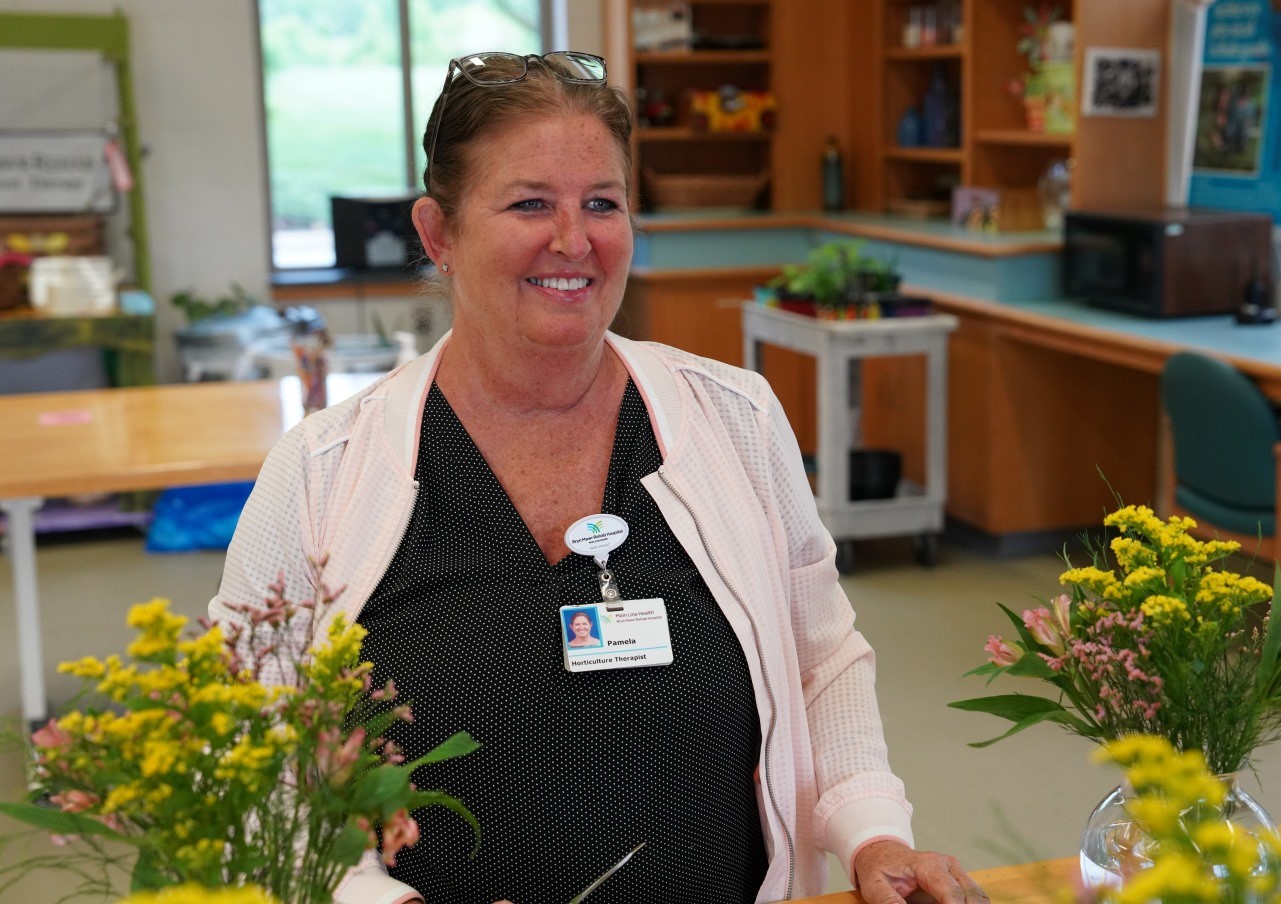 A woman with a name badge smiles while arranging flowers in a bright room with tables and shelves, creating a welcoming and cheerful atmosphere.