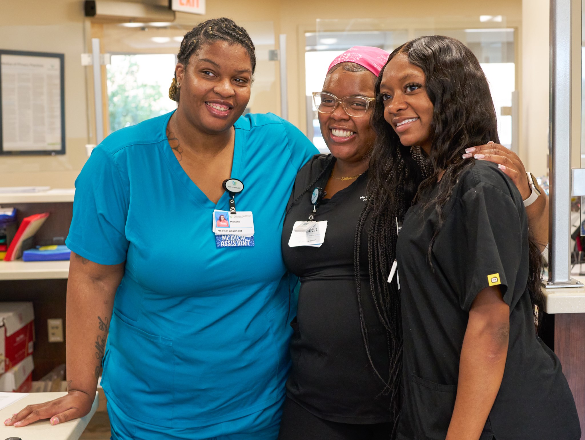 Three women in medical uniforms smile and pose together inside a healthcare facility, showing camaraderie and teamwork. One wears blue scrubs, another black, and the third has a pink headband and glasses.