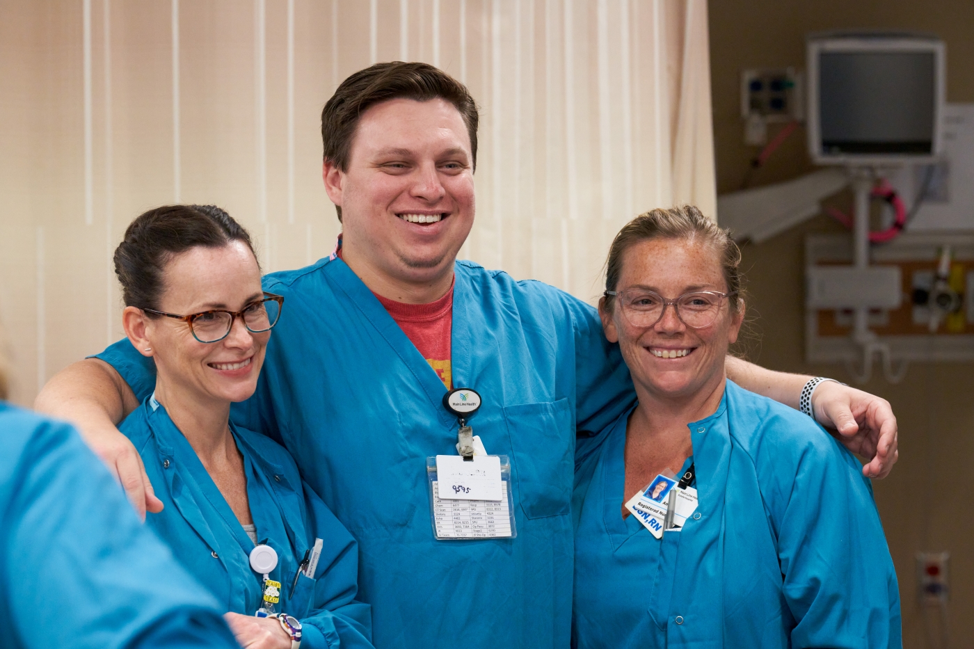 Three healthcare workers wearing blue scrubs and name badges stand together, smiling, in a hospital setting. Two women are on either side of a man, and a medical curtain is visible in the background.