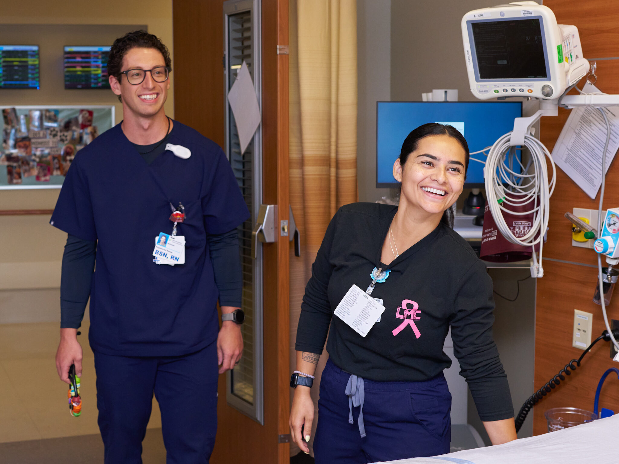 Two healthcare professionals, a man and a woman, smile while standing beside a hospital bed in a patient room equipped with medical monitors and equipment.