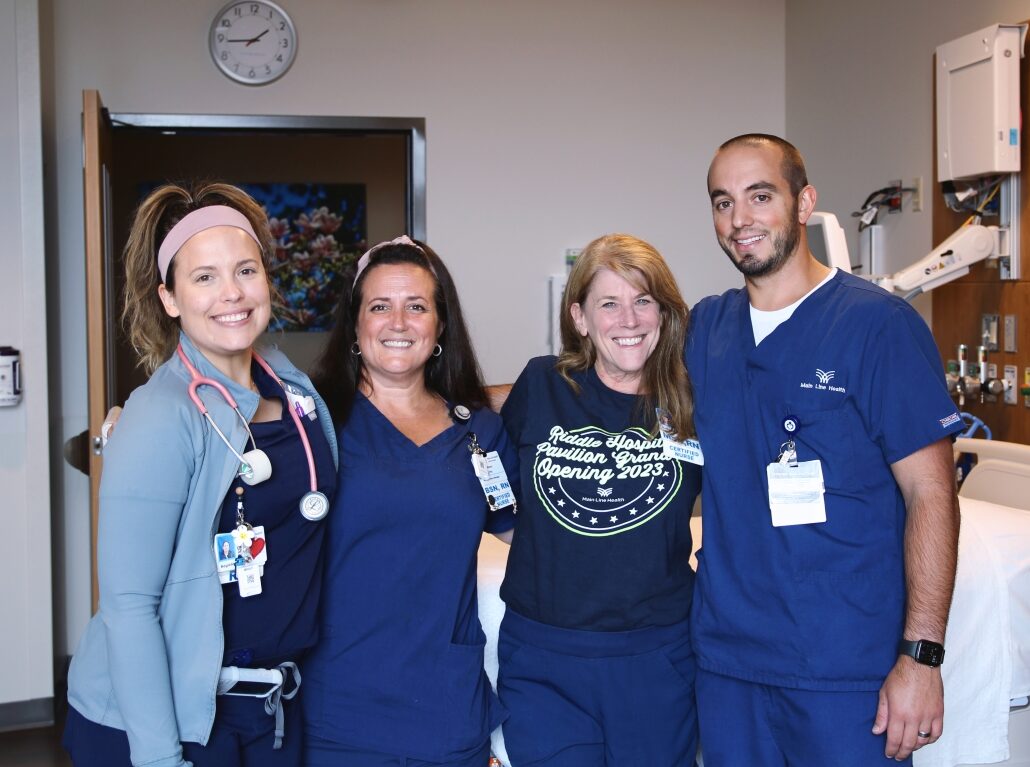 Four smiling healthcare workers pose together in a hospital room, with medical equipment and a hospital bed in the background. Three wear navy blue scrubs and one wears a T-shirt that says "Nursing 2023.