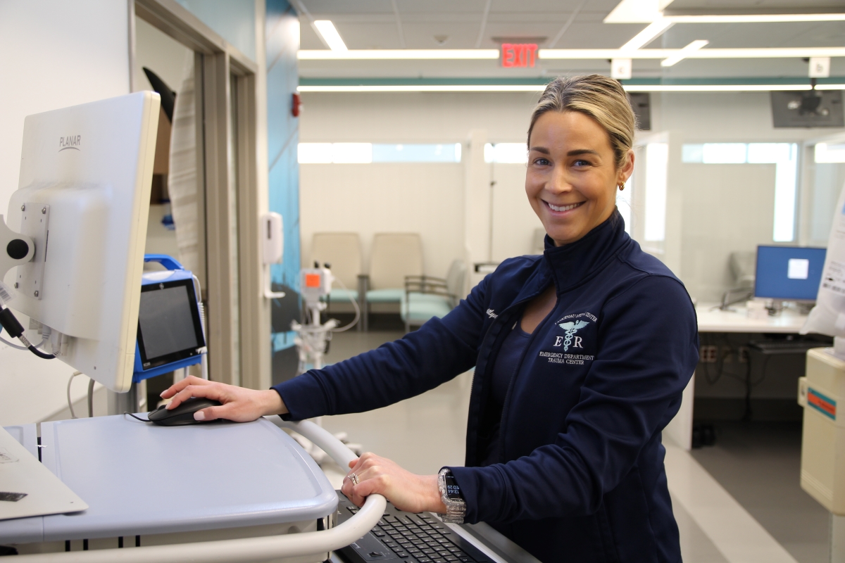 A woman in medical scrubs and a dark jacket stands at a computer workstation in a bright hospital or clinic, smiling at the camera. Examination chairs and medical equipment are visible in the background.
