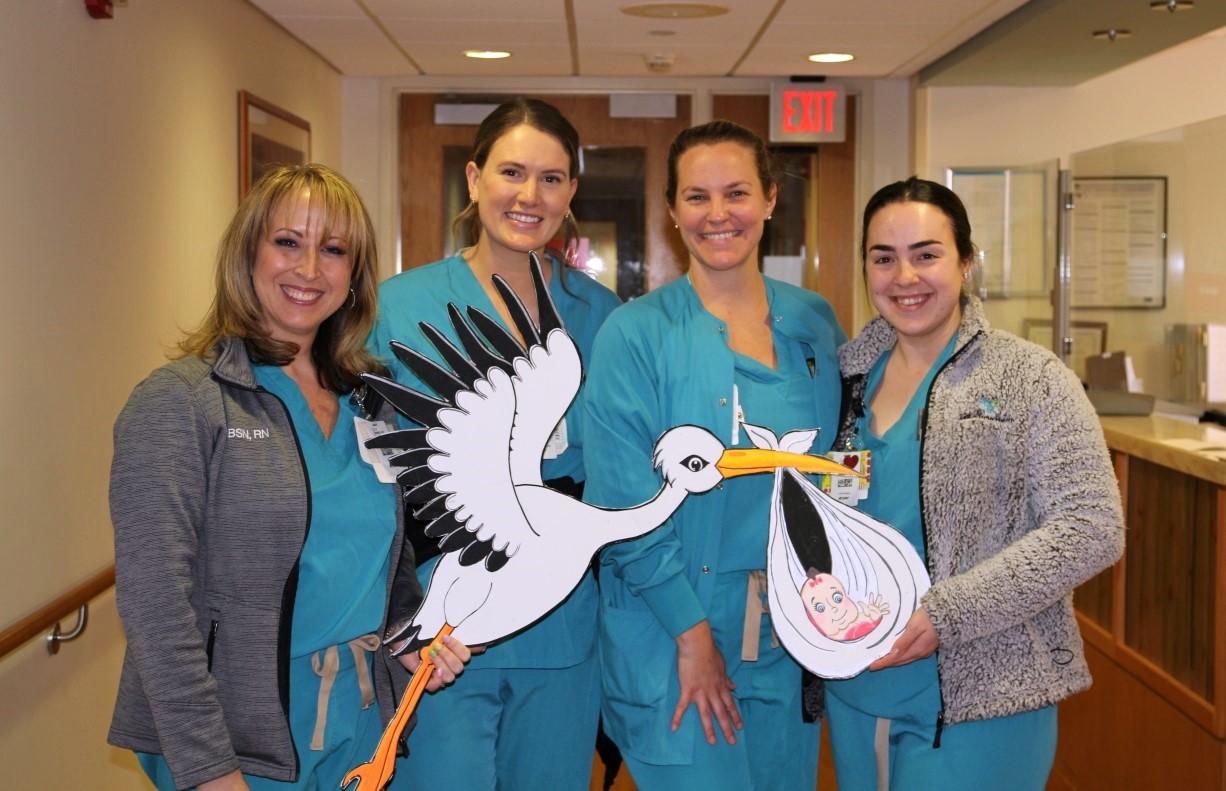 Four smiling women in teal scrubs stand in a hospital hallway, holding large cutouts of a stork carrying a baby. One woman is in a gray jacket, and the group appears cheerful and friendly.