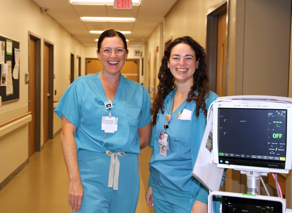 Two women wearing blue medical scrubs and ID badges stand smiling in a hospital hallway next to medical equipment. The hallway features bulletin boards and fluorescent lighting.