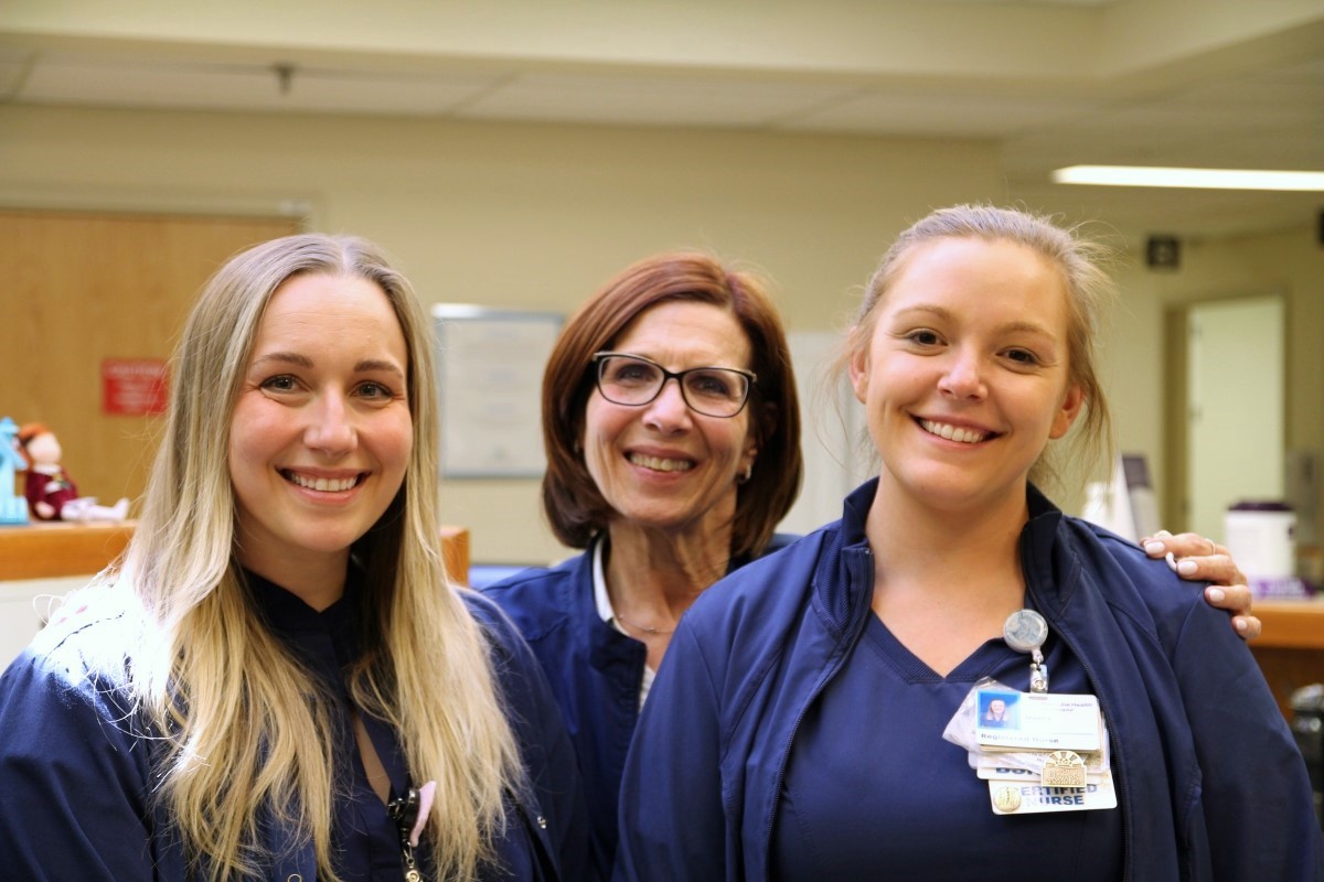 Three women in navy blue scrubs, smiling and standing together in what appears to be a hospital or medical facility. Two have blonde hair and one has brown hair and glasses. They are wearing name badges.