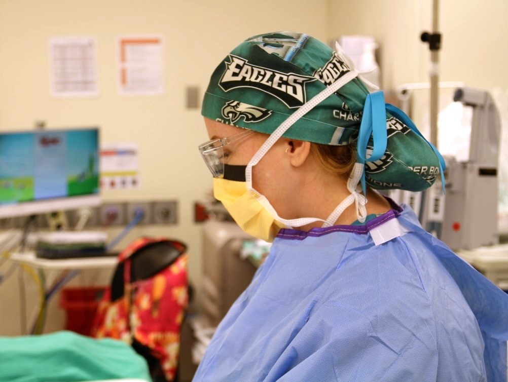 A healthcare worker wearing a Philadelphia Eagles cap, protective goggles, a yellow face mask, and blue scrubs stands in a medical setting with monitors and equipment in the background.