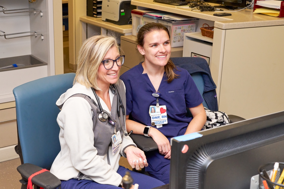 Two nurses in scrubs sit side by side at a desk, smiling while looking at a computer screen. One nurse has a stethoscope around her neck; both wear ID badges and appear to be engaged in a positive conversation or meeting.