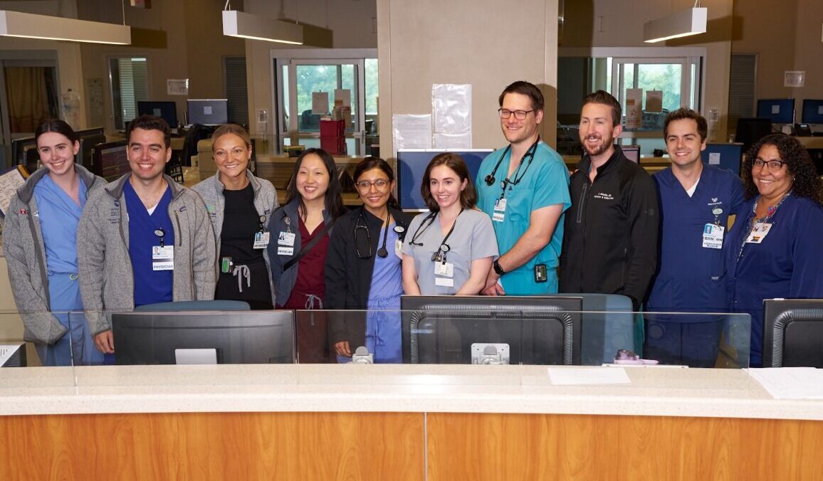 A group of ten healthcare professionals, dressed in scrubs and lab coats, stand and smile together behind a reception desk in a hospital or clinic setting. Computer monitors and medical equipment are visible.