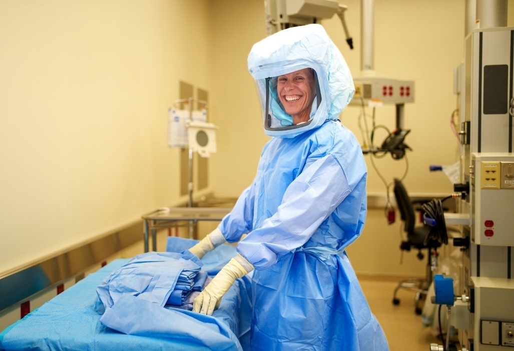 A person wearing full protective medical gear and a powered air-purifying respirator smiles while standing in a hospital room, preparing surgical drapes on a table.
