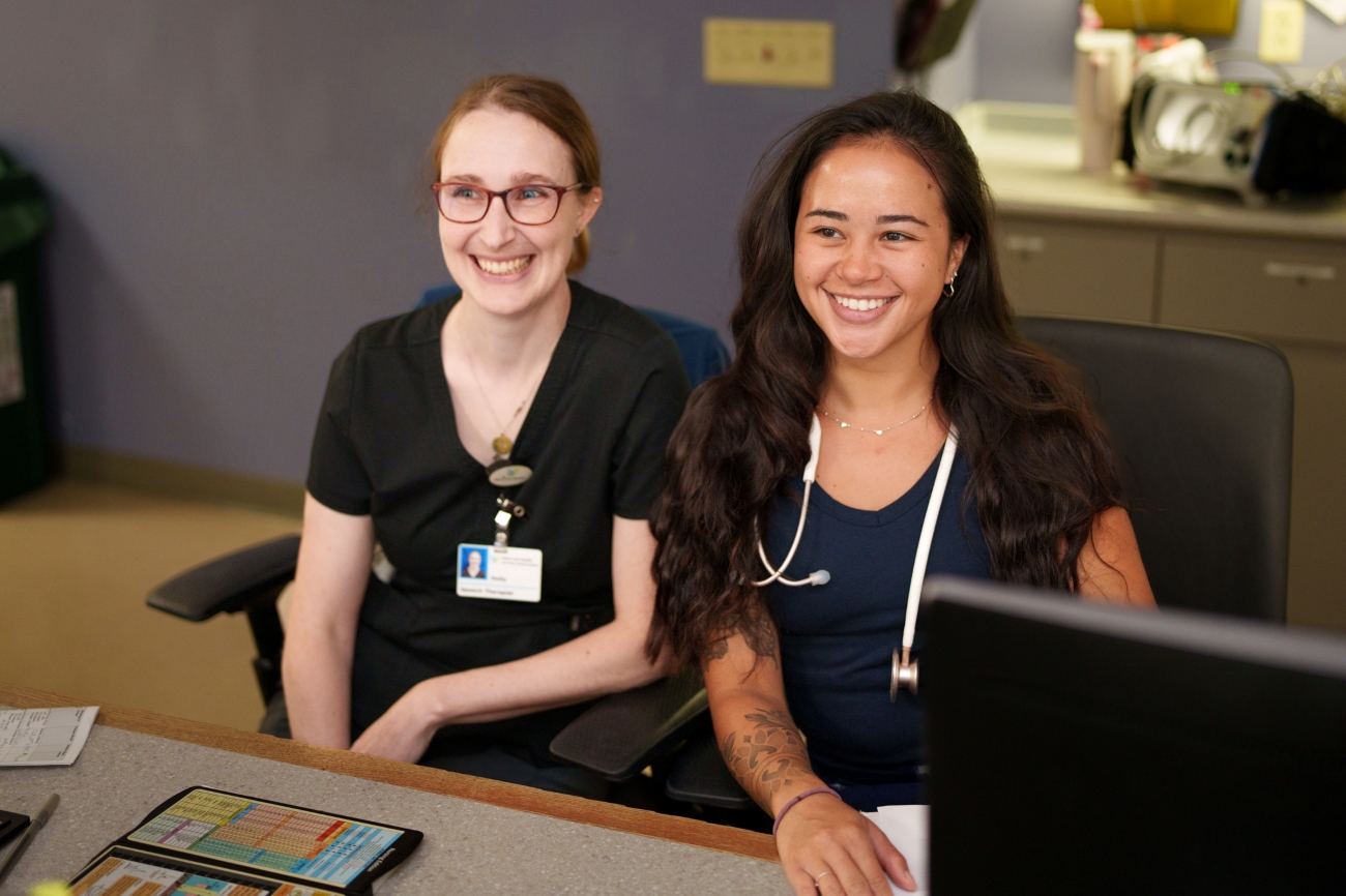 Two women in medical uniforms, one with a stethoscope, sit at a desk smiling. A tablet and notepad are on the desk, and a computer monitor is in front of them in a clinical office setting.